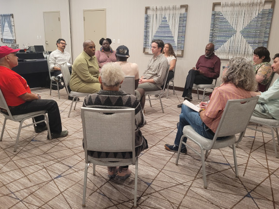 Twelve people sitting in a circle on being metal chairs in a beige conference room.