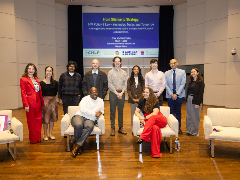 A group of 9 peopel standing, with two seated in front of an info screen on a conference room stage.