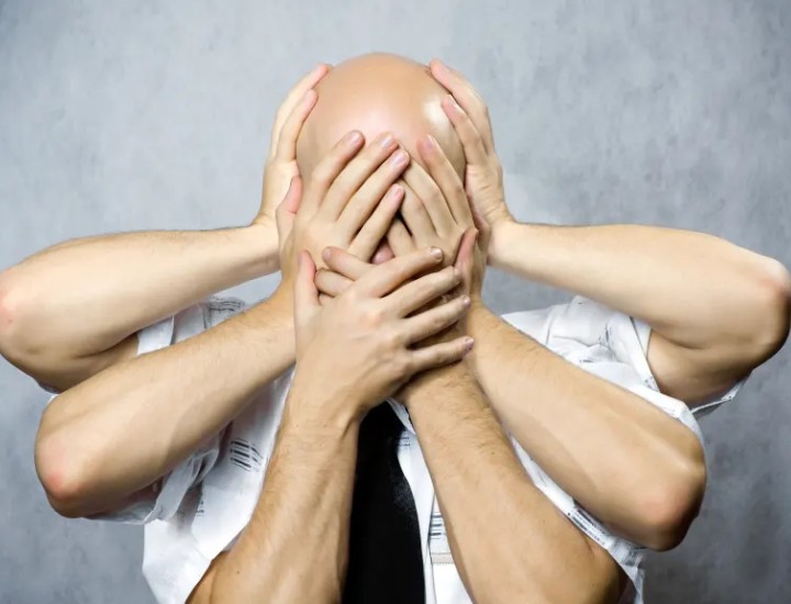 A bald, white man facing the camera with three pairs of white hands covering his face.