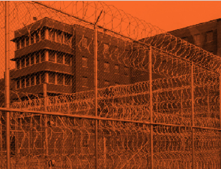 Black and orange duotone of a brick prison building behind barbed wire
