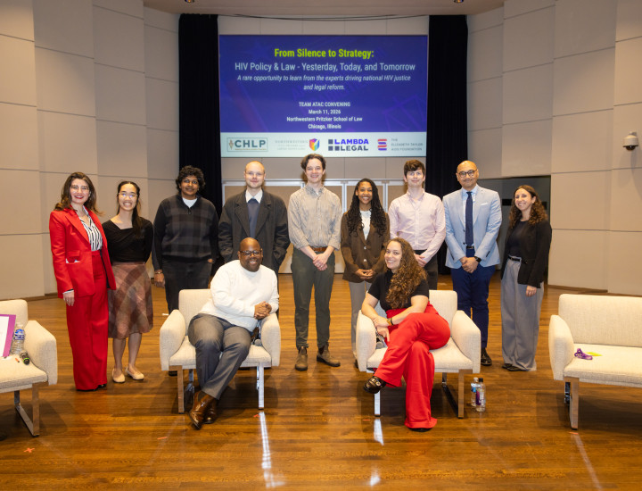 A group of 9 peopel standing, with two seated in front of an info screen on a conference room stage.